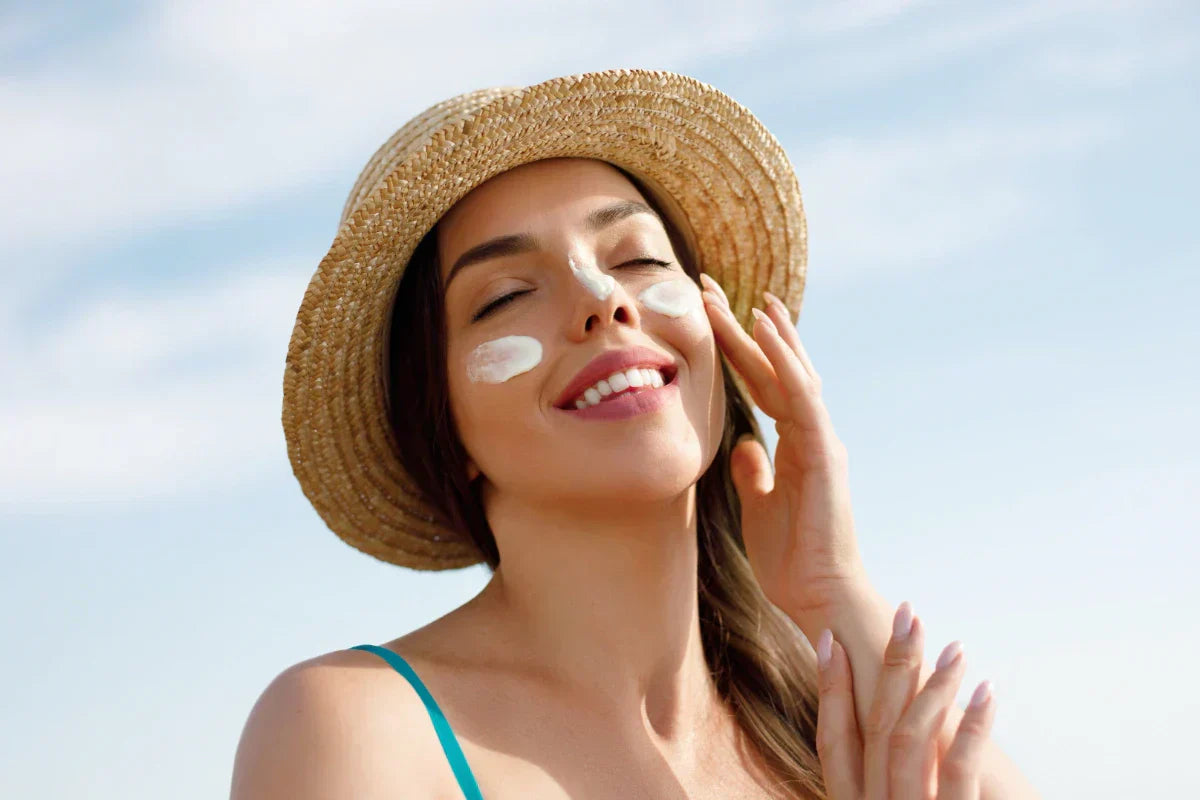 Woman wearing a straw hat and applying sunscreen to her cheeks and nose while smiling under a sunny sky, promoting daily sun protection with niacinamide.
